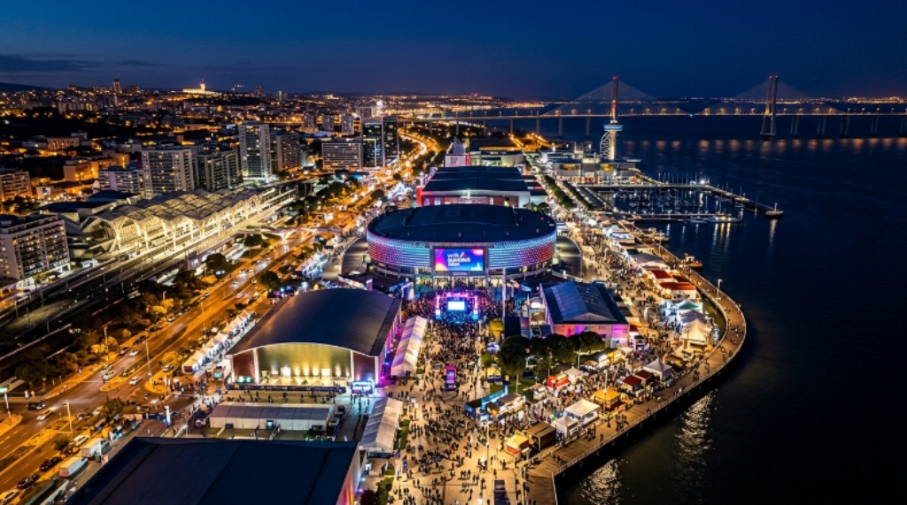 Aerial view of Parque das Nações in Lisbon at night during Web Summit 2026, showing the illuminated Altice Arena and the waterfront district